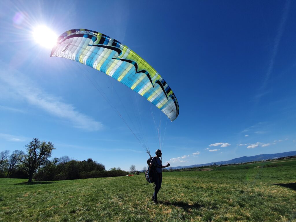 Person on the ground with paraglider in the air on a grass field with sun shining