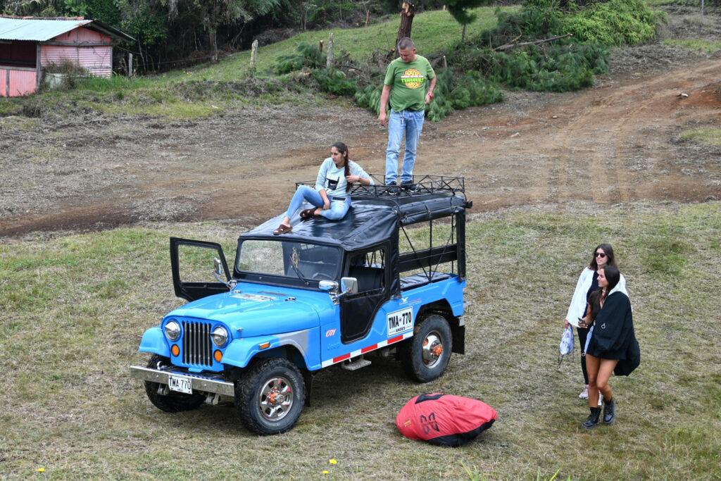 Blue jeep with people sitting and standing on the roof, unloading paragliding equipment