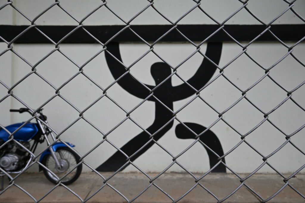 Street art: black figure on a wall lifting a black bar with a motorcylce parked in front (photo taken through a fence)