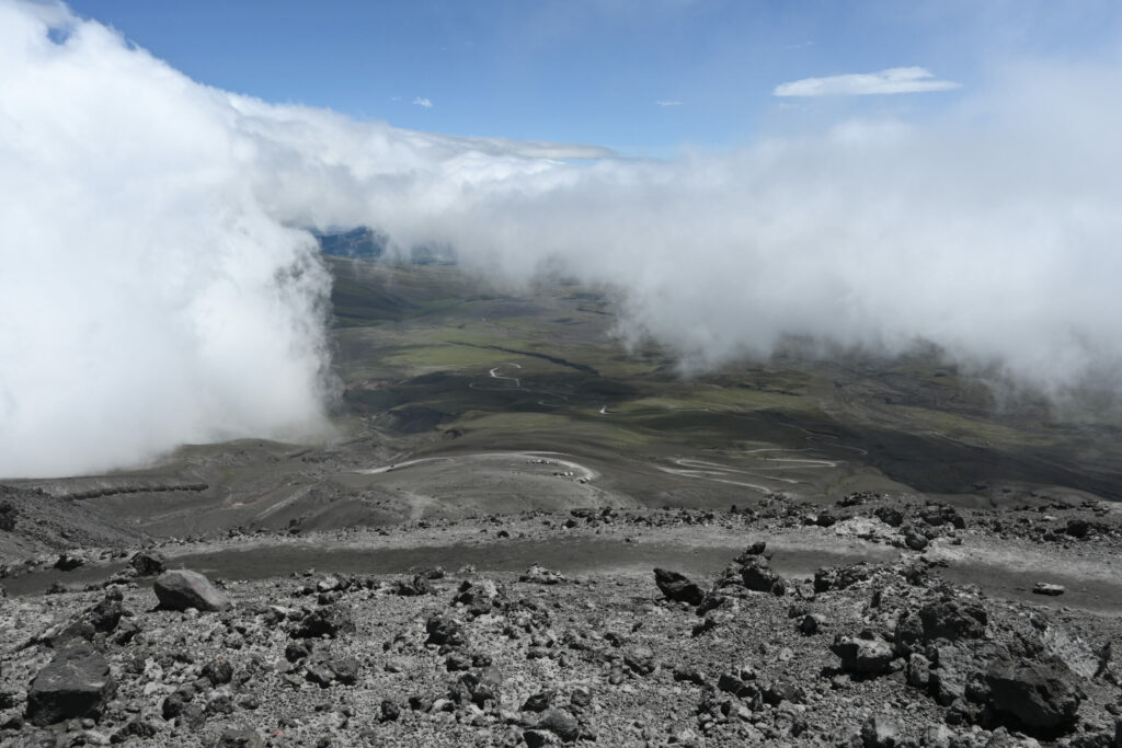 Picture from a mountain down to the flats. Rocks in the foreground, green land in the background and clouds obscuring a bit of the view