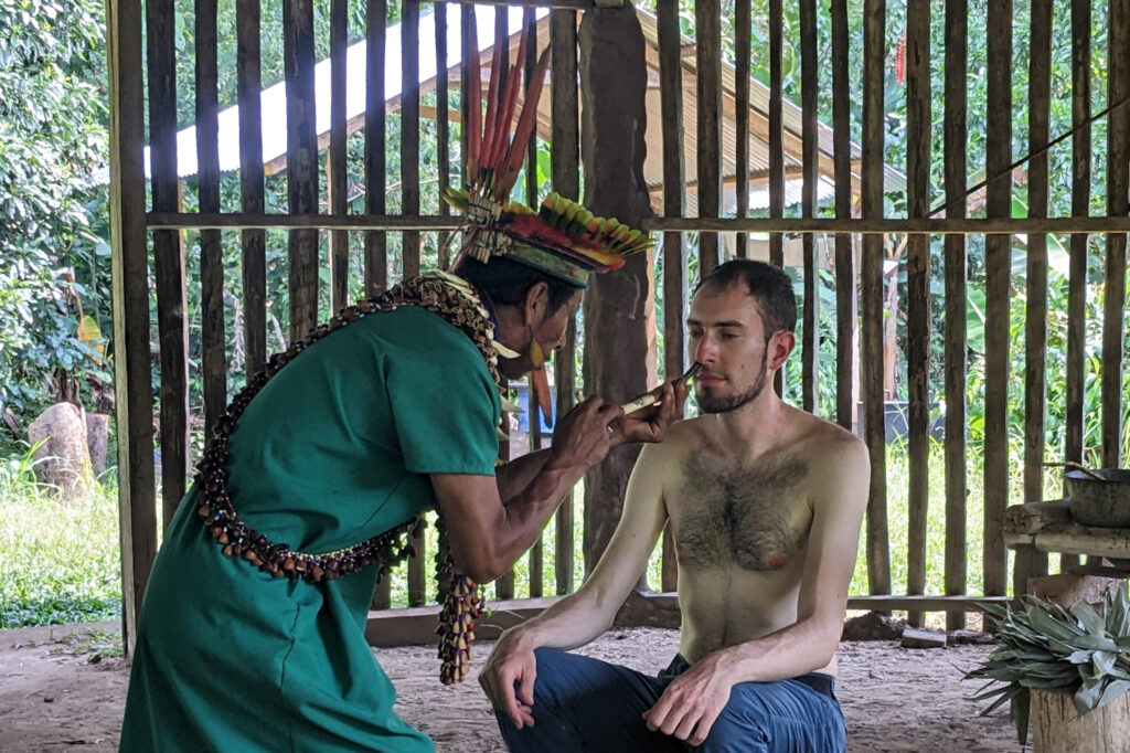 Person sitting with bare top getting tobacco blown into the nose by a Shaman with a green custome and feathers attached to the head